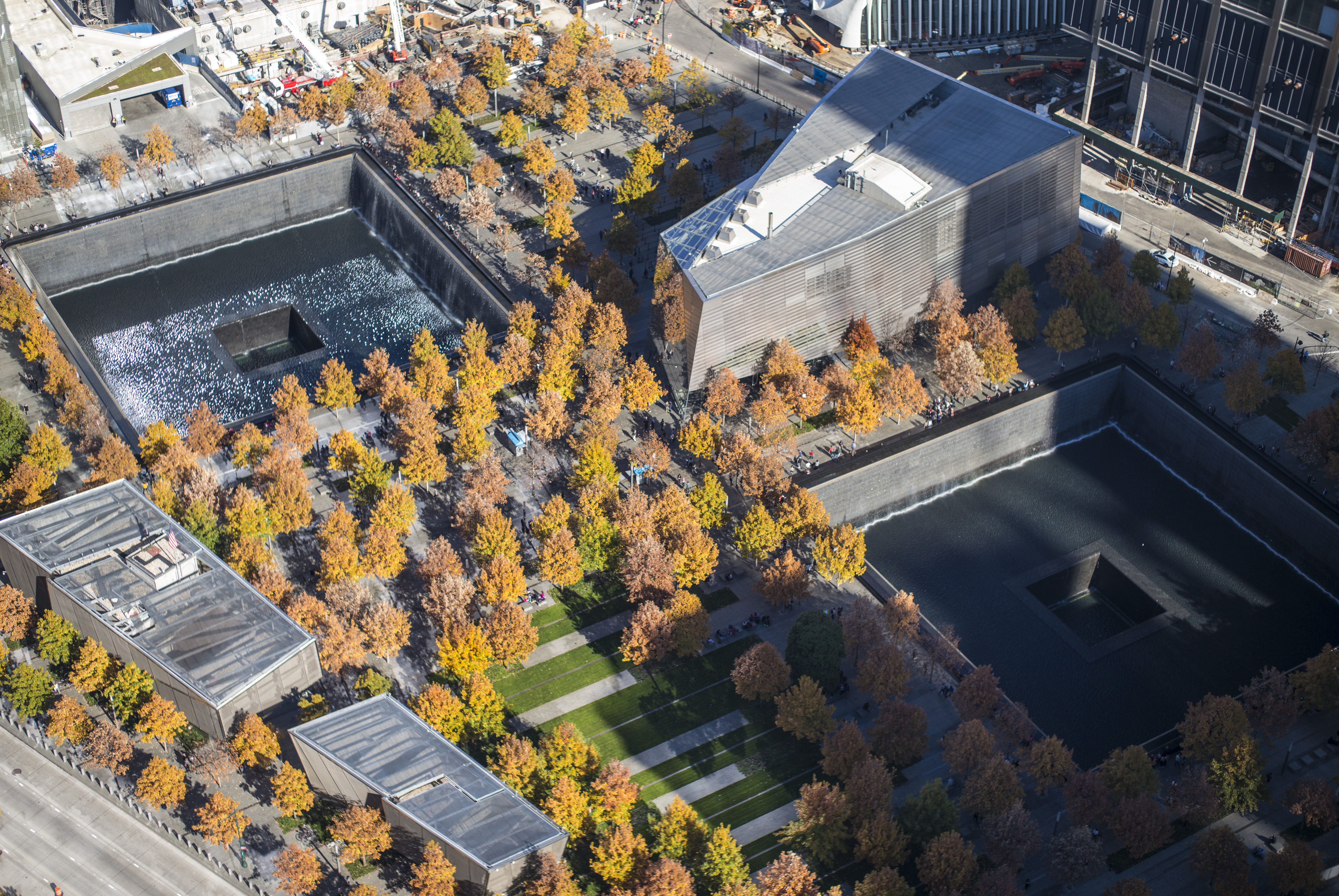 An aerial photo of the 9/11 Memorial on a fall day shows the colorful yellow and orange leaves on trees planted throughout the plaza. Shadows and sunlight fall across the reflecting pools and buildings of the Memorial.