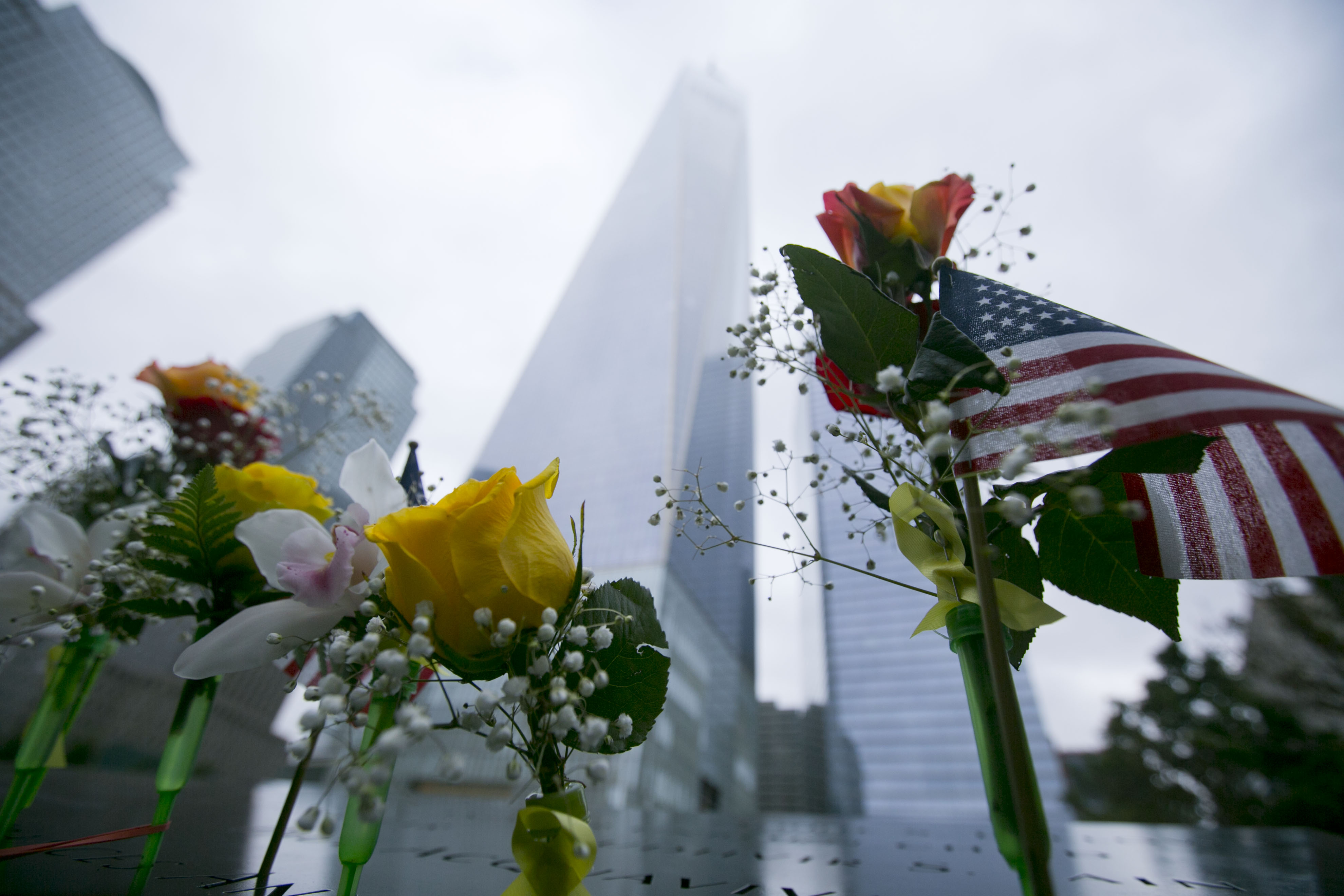A small American flag and flowers, including yellow roses, stand at names on a bronze parapet at the 9/11 Memorial. One World Trade Center towers above on an overcast day.