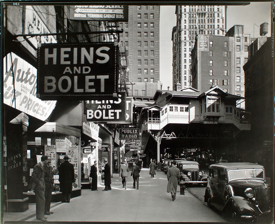 A view shows “Radio Row” in Manhattan in 1936. Signs line storefronts. An elevated subway station is seen in the background.