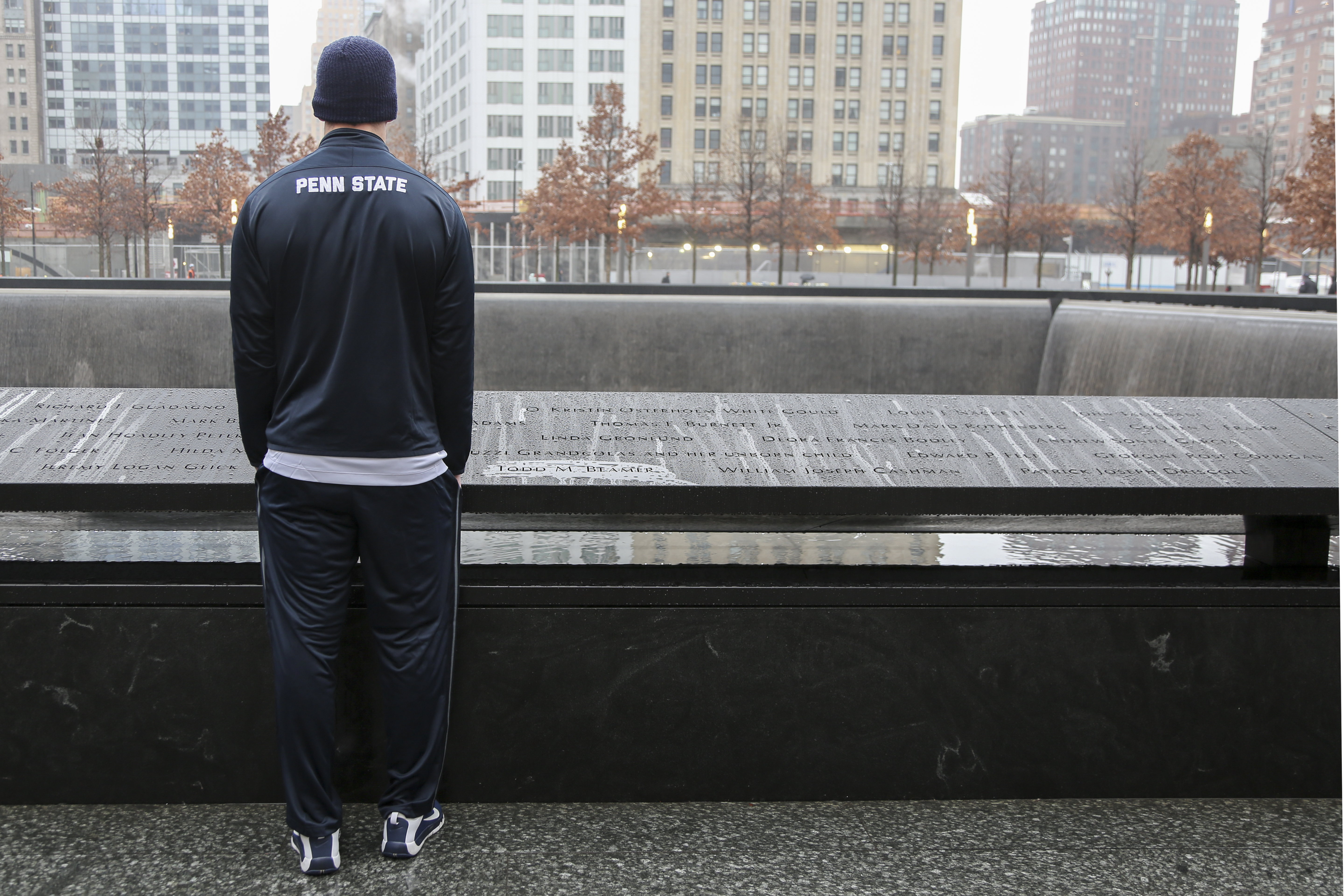 A Penn State University athlete honors victims of 9/11 at the Memorial. He is standing beside a bronze parapet with victims names on a rainy day. He is wearing a blue tracksuit that reads “Penn State.”