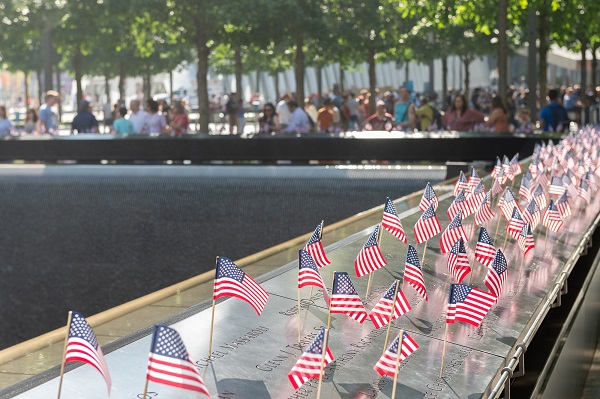 Dozens of small American flags have been placed at the names of victims at the 9/11 Memorial on a sunny day. Visitors tour Memorial plaza in the background.