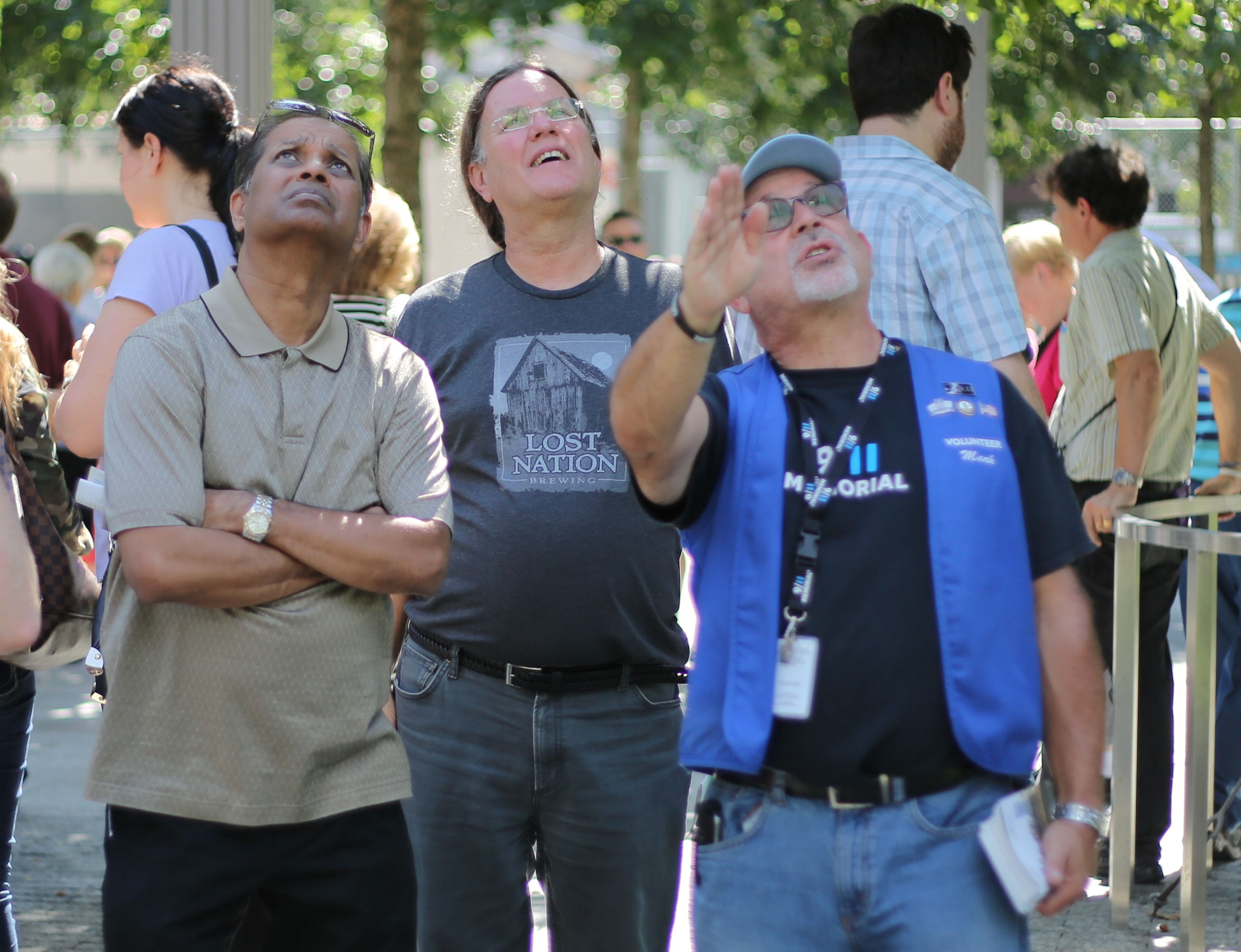 A tour guide wearing a blue vest and a black lanyard points his right arm at an object out of view. Two men beside him look up at the object.