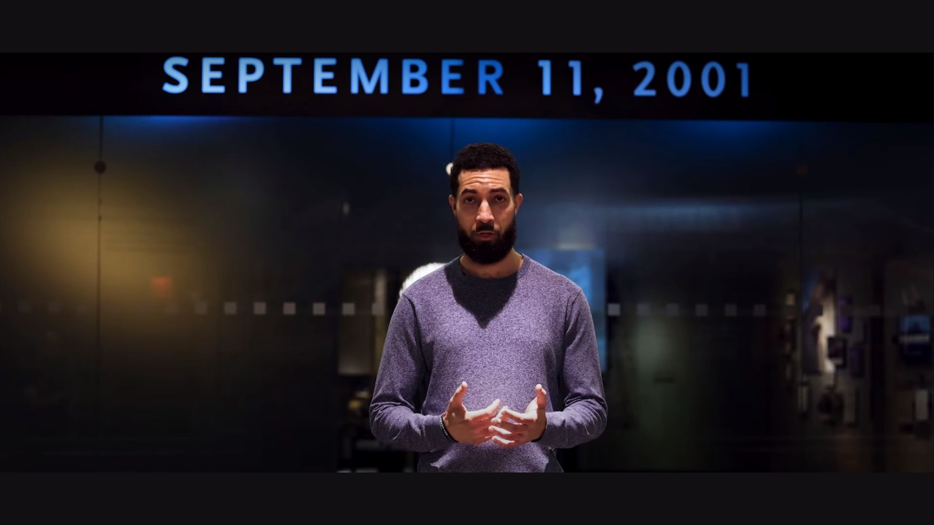 A man with dark hair and a beard, wearing a medium blue sweater, speaks in front of a darkened background. At the top, the date September 11, 2001 is visible in text.