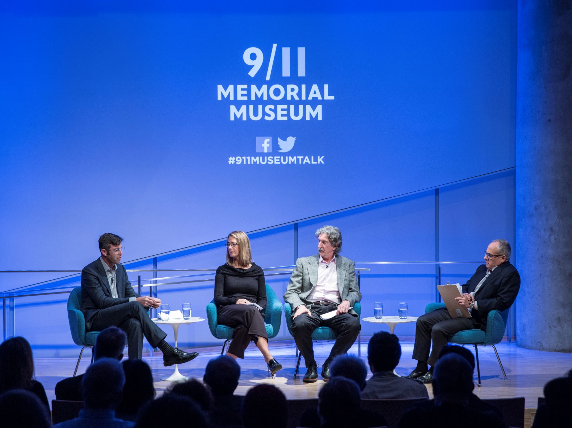 Handel Architects LLP partner Michael Arad, Paul Murdoch Architects President Paul Murdoch, and KBAS partner Julie Beckman are onstage as they take part in the public program, The Architecture of Remembrance. Clifford Chanin, the executive vice president and deputy director for museum programs, is seated to their left holding clipboard. The 9/11 Memorial & Museum logo is projected above the four of them. The silhouettes of audience members are in the foreground.