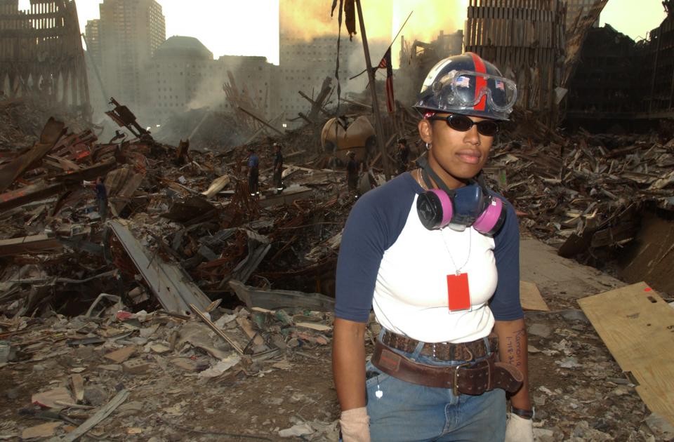 Ramona Diaz-Allegrini poses for a photo in front of a smoky pile of debris at Ground Zero. The Koenig Sphere and the steel facades of the Twin Towers are visible in the background.