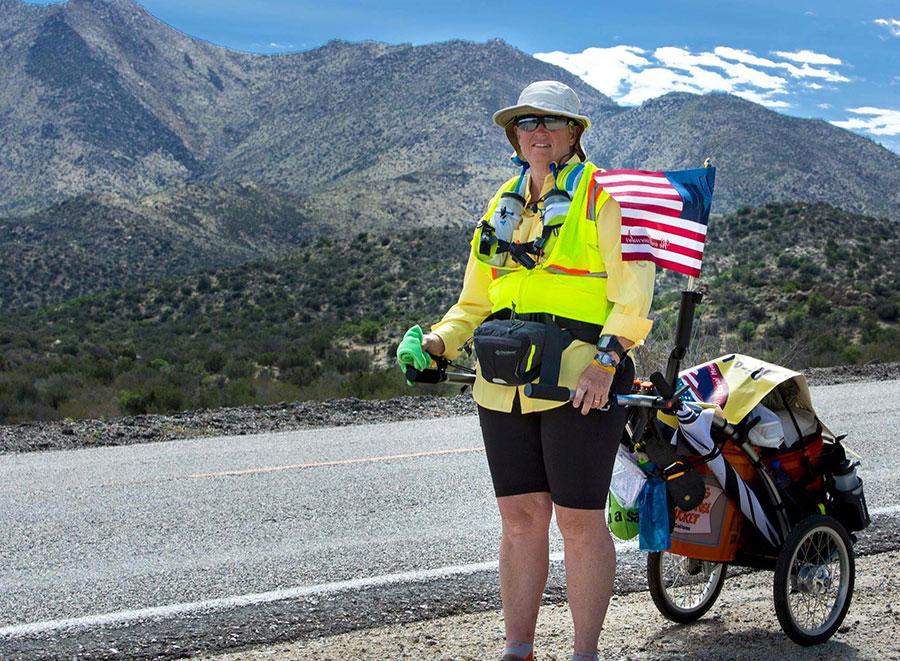 Jan Walker poses for a photo in front of a mountain range during her four-month journey across the U.S. She is standing beside a road as she wheels her belongings in a cart behind her.