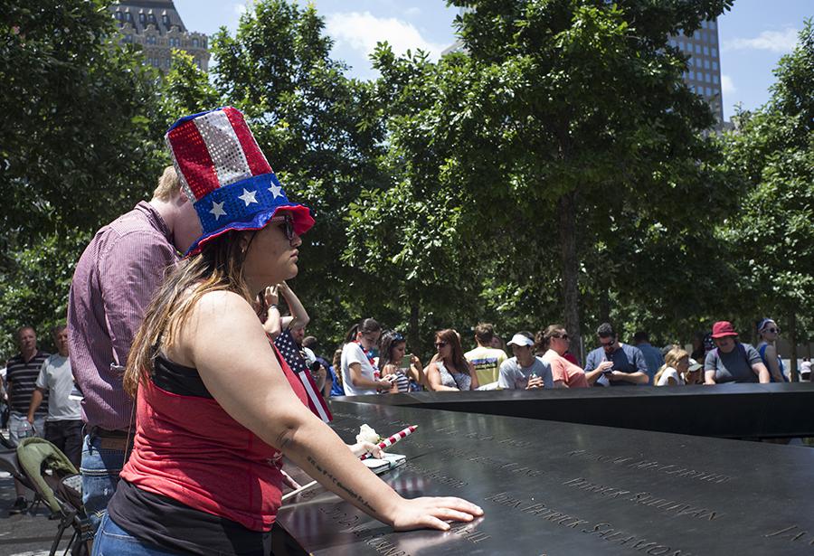 A woman in a stars and stripes tophat puts her arms on a bronze parapet at the Memorial as she looks out over a reflecting pool. Other visitors are gathered behind her.
