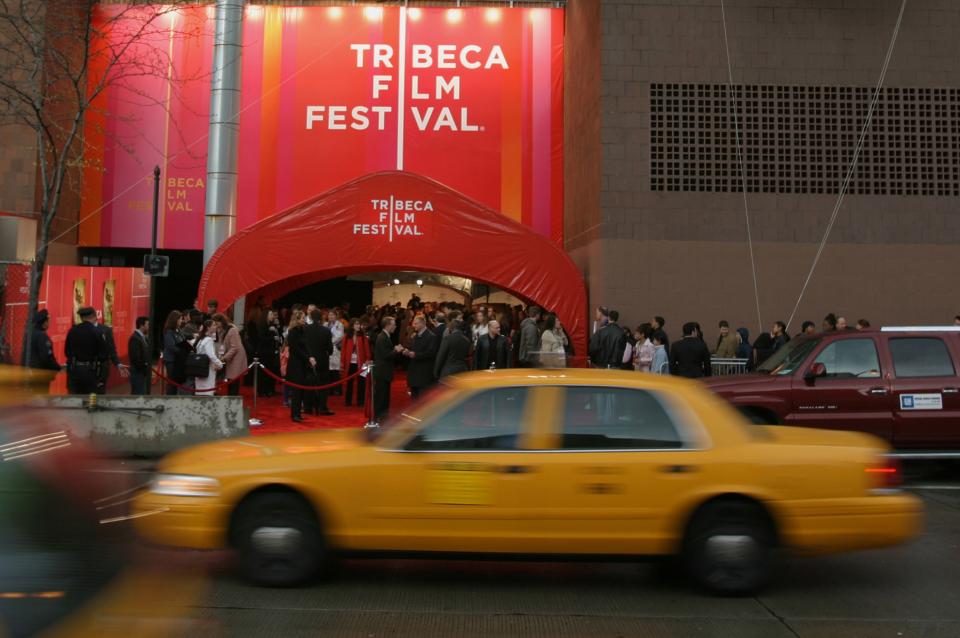 A yellow taxi cab passes the entrance to the Tribeca Film Festival. A crowd of people is under an illuminated red sign that reads “Tribeca Film Festival.”