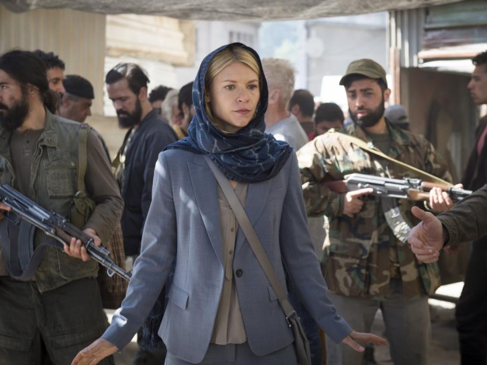 Claire Danes portrays Carrie Mathison in a scene from “Homeland.” She is wearing a blue suit and veil and is surrounded by men in camouflage holding guns.