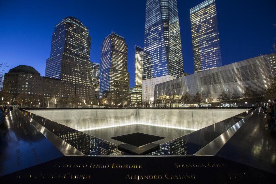 One World Trade Center towers over the south pool of the 9/11 Memorial on a clear night. Water cascades down the illuminated reflecting pool.
