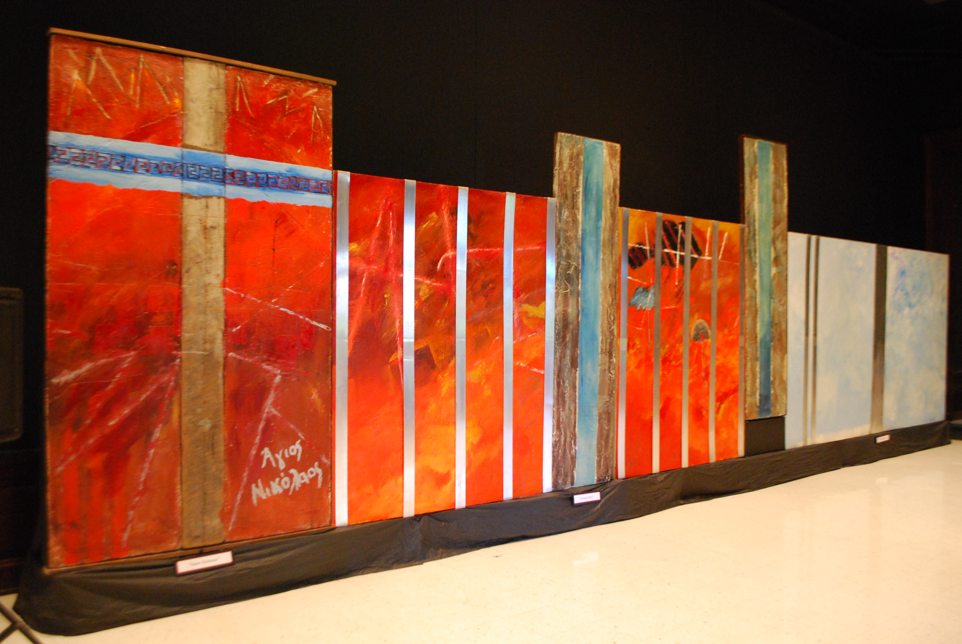 Long interconnected work with (l. to r.) Large wooden beam with crossing Greek key motif surrounded by brilliant red;  fiery red canvas panels with smaller forms, overlain by shiny metal laths; and two large abstract sections with white-obscured sky blue, overlain with metal laths.