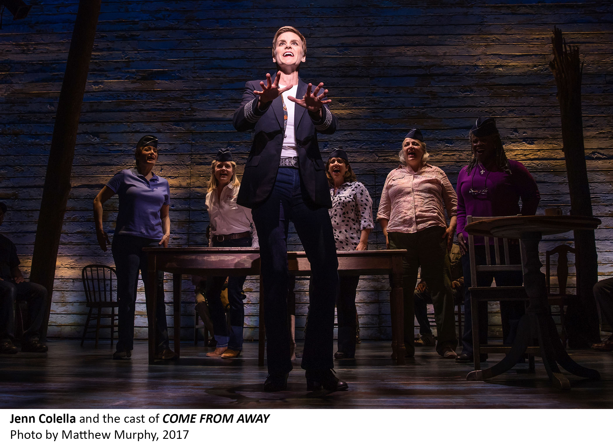Jenn Colella and several other members of the cast of Come From Away perform onstage. Colella is gesturing as a spotlight shines on her.