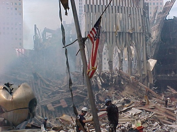 The smashed Koenig Sphere and steel tridents from the tubular framework of the Twin Towers stand amid piles of debris at Ground Zero. An American flag has been raised on a pole sticking up from the debris. Two men are leaning against the pole.