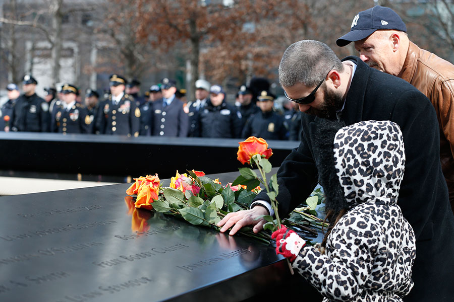 A young girl in a cheetah patterned jacket places yellow and red roses at a name on the Memorial during the ceremony commemorating the 23rd anniversary of the World Trade Center bombing. A man who is standing next to her is placing his hand on a bouquet of roses that have been placed nearby.