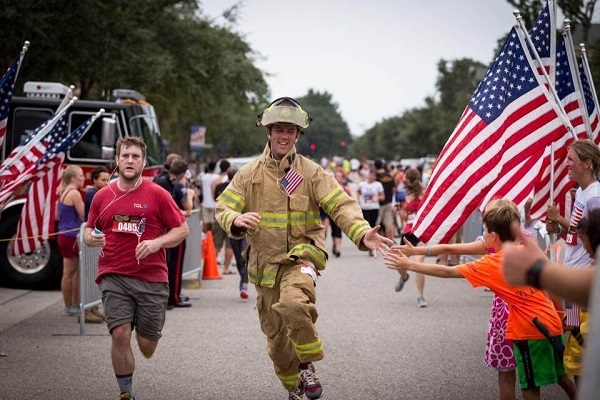 A smiling firefighter in bunker gear gives a boy and girl a high five as he runs in an event. American flags and spectators line the route as other runners follow behind him.
