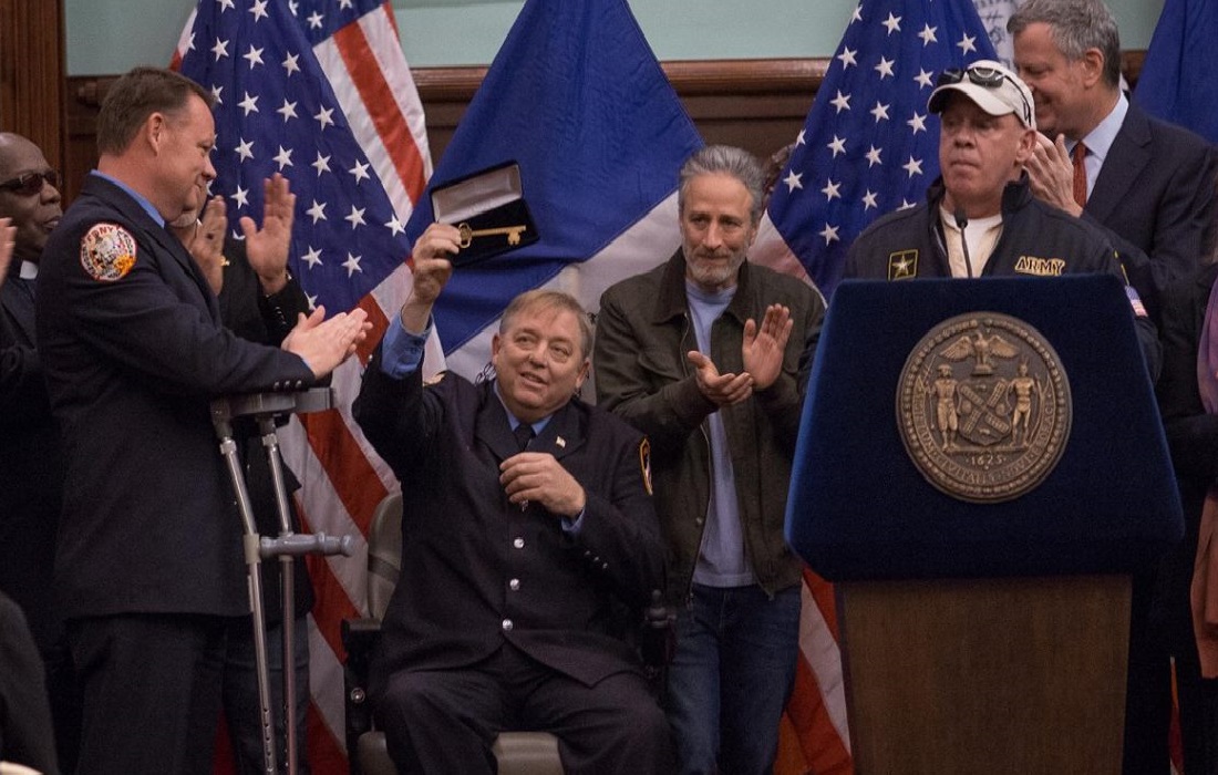 Former FDNY firefighter Ray Pfeifer holds up the key to New York City that was presented to him by Mayor Bill de Blasio at City Hall. Comedian John Stewart claps as he stands beside Pfiefer.