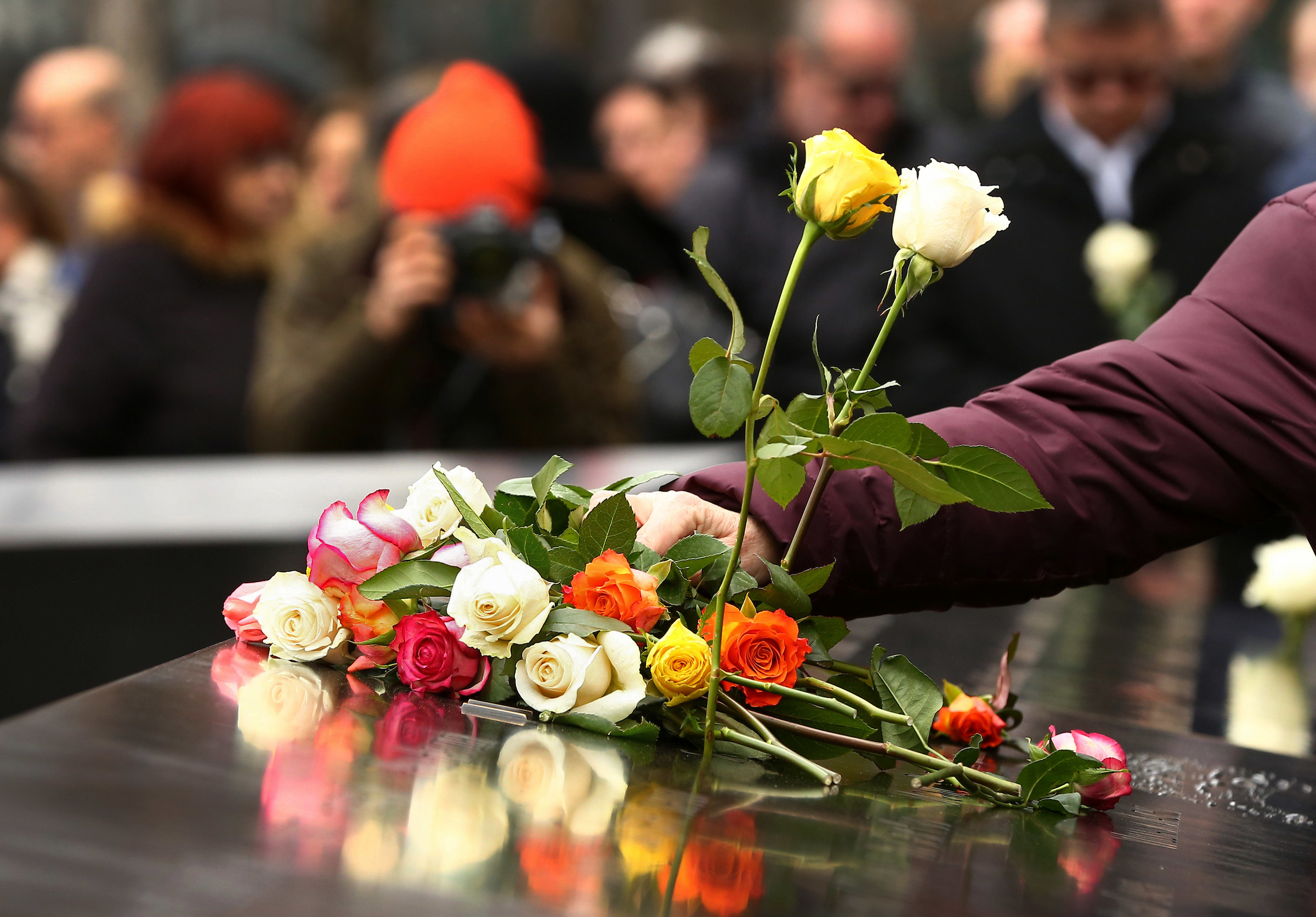 A person’s arm is seen placing a bouquet of red, orange, yellow, and whiite roses on a bronze parapet at the Memorial.