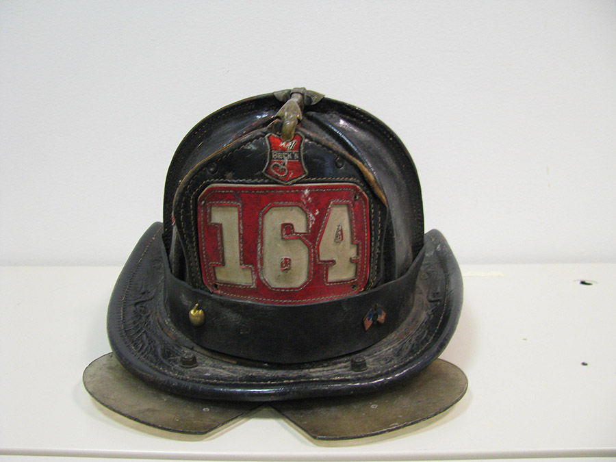 Robert Beckwith’s black and red fire helmet is displayed on a white surface at the Museum. The helmet features features the number 164.