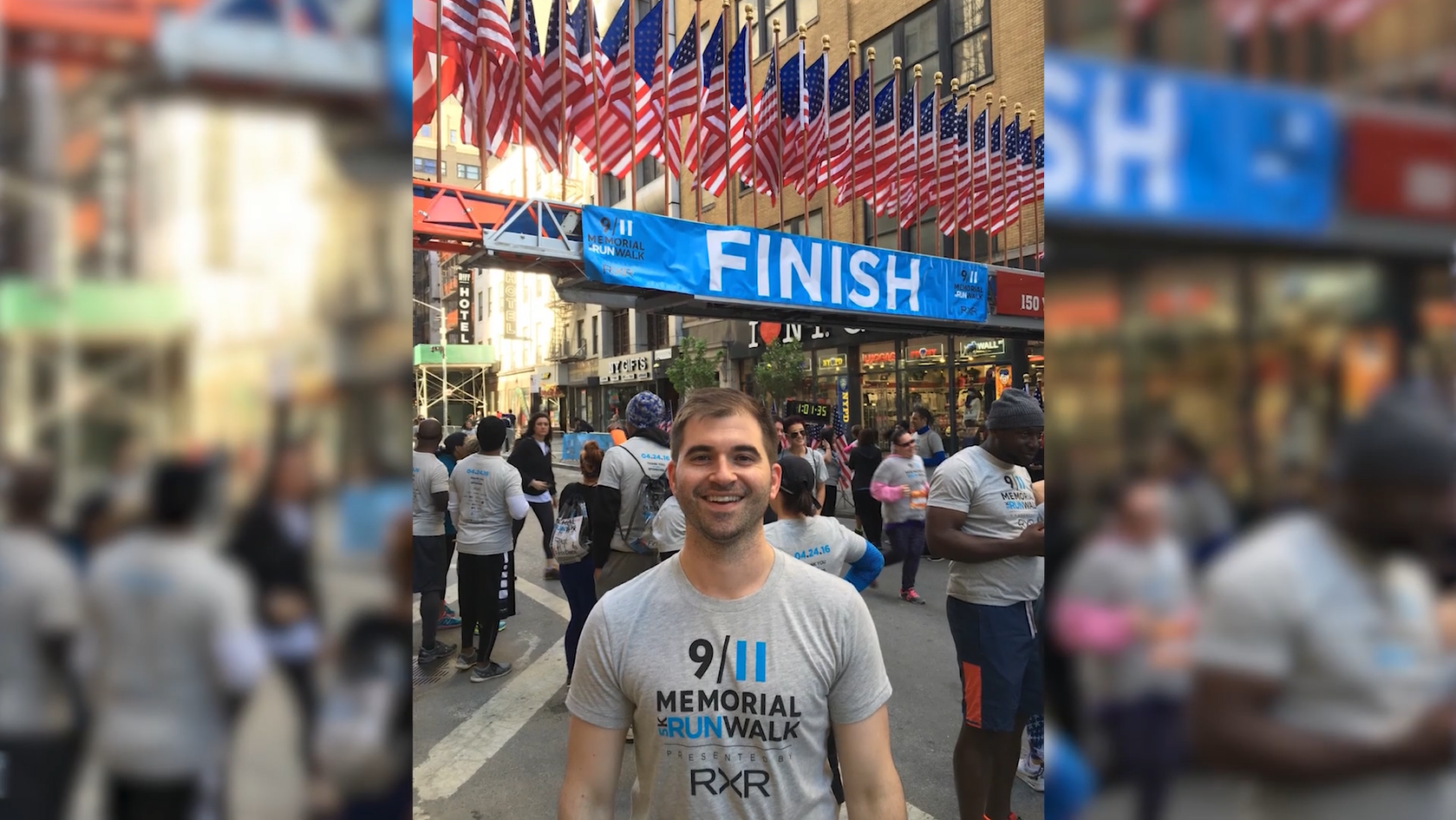 Alex Zablocki, a Museum member and 9/11 Memorial Run/Walk participant, smiles at the finish line in 2016. Other participants cross the finish line behind him. Dozens of American flags stand above the finish line sign.