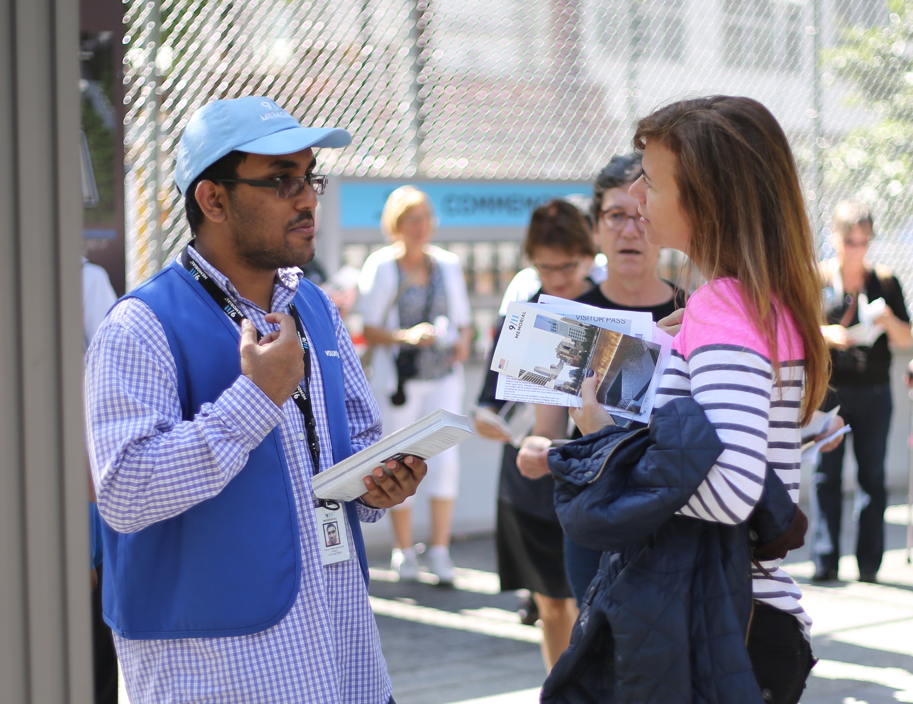 A 9/11 Memorial Museum volunteer speaks with a woman on the Memorial plaza. The volunteer is wearing a blue vest and a blue hat and grasping an informational booklet.