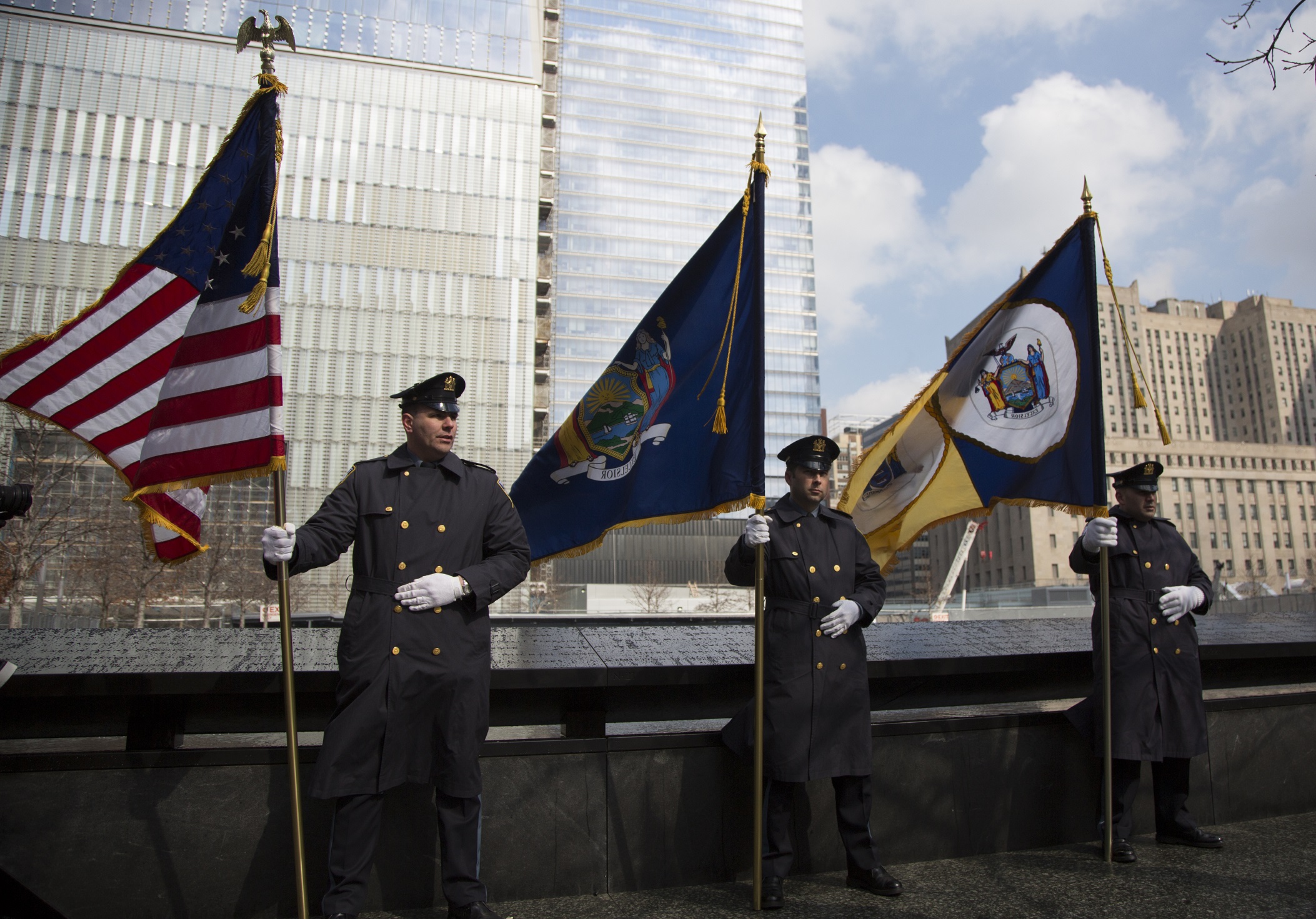 Three police officers in formal uniforms hold large flags, including an American flag and a New York State flag, as they stand beside a reflecting pool on Memorial plaza.