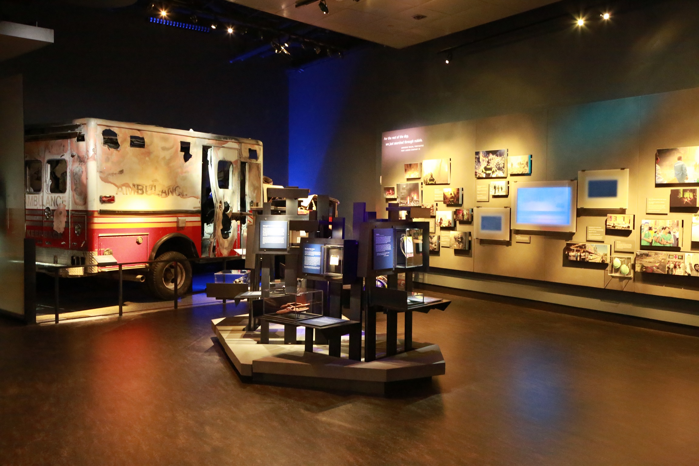 A section of the Museum’s Historical Exhibition is seen without any visitors. A damaged FDNY ambulance sits off to the right, while various images and interactive screens are on a wall to the right.