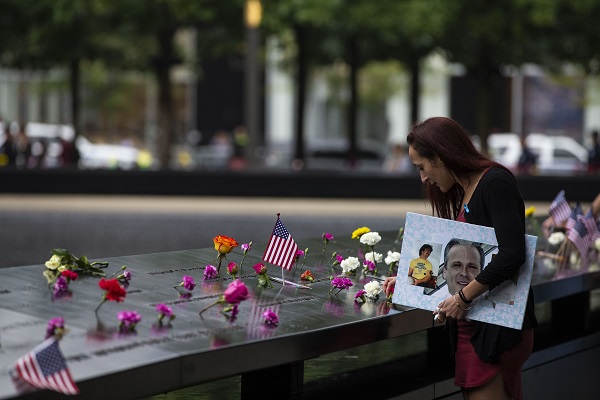 A woman wearing a blue ribbon stands beside a bronze parapet on the 9/11 Memorial. She is touching a name on the Memorial while holding a cardboard sign with photos of a man. Dozens of flowers of various colors have been placed at names around here. Small American flags have also been placed at the names.