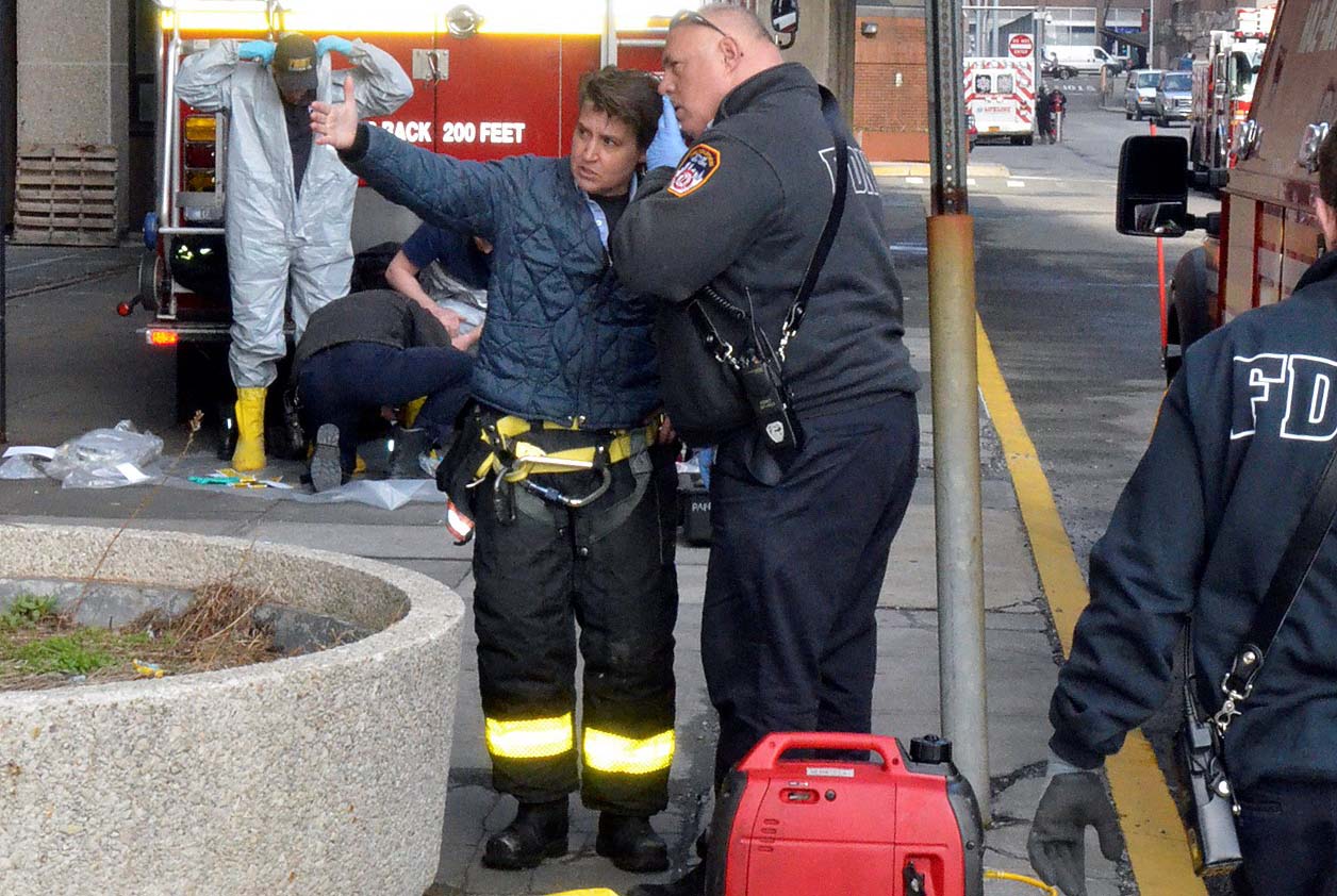 FDNY Lt. Adrienne Walsh wears bunker gear as she speaks to a man in an FDNY jacket while standing on the street and pointing at something out of view.