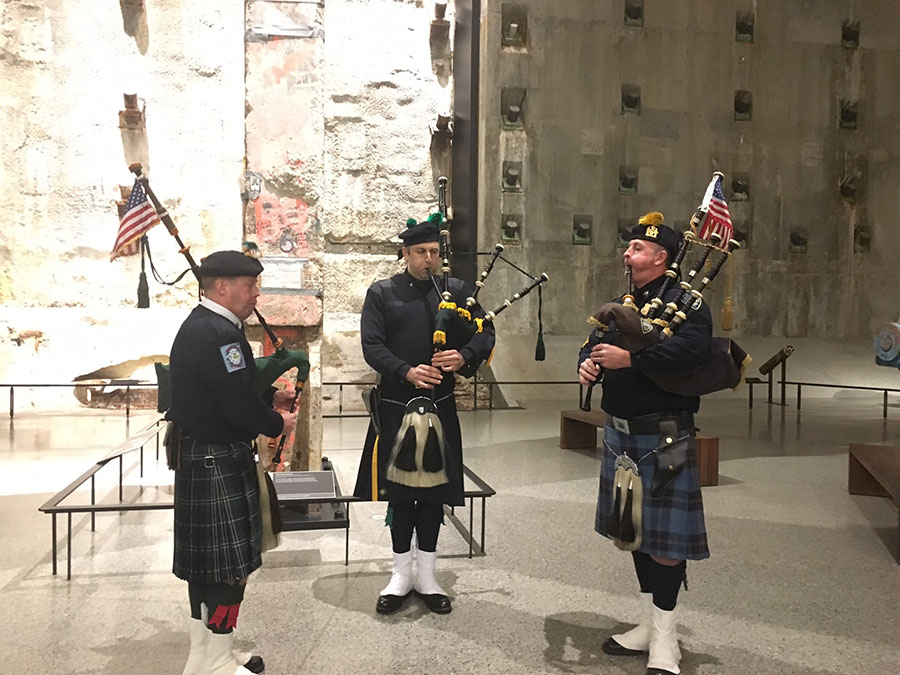 FDNY officer Bob King, NYPD officer Eamon Nugent, and PAPD officer Brian Ahern participate in a weekly bagpipe tribute in Foundation Hall. They are wearing traditional outfits, including kilts. The slurry wall is lit up behind them.