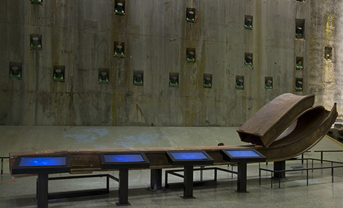A large rusty beam from the Twin Towers is displayed beside four signing screens in Foundation Hall. The concrete slurry wall is visible in the background.