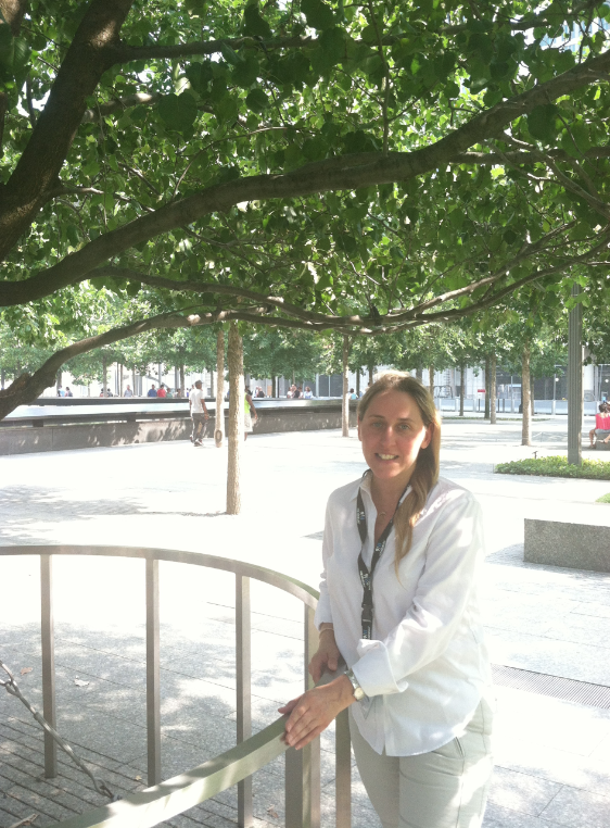Volunteer Catherine Nadal stands in the shade of the Survivor Tree on the 9/11 Memorial plaza. Visitors to the Memorial can be seen beside a reflecting pool behind her.
