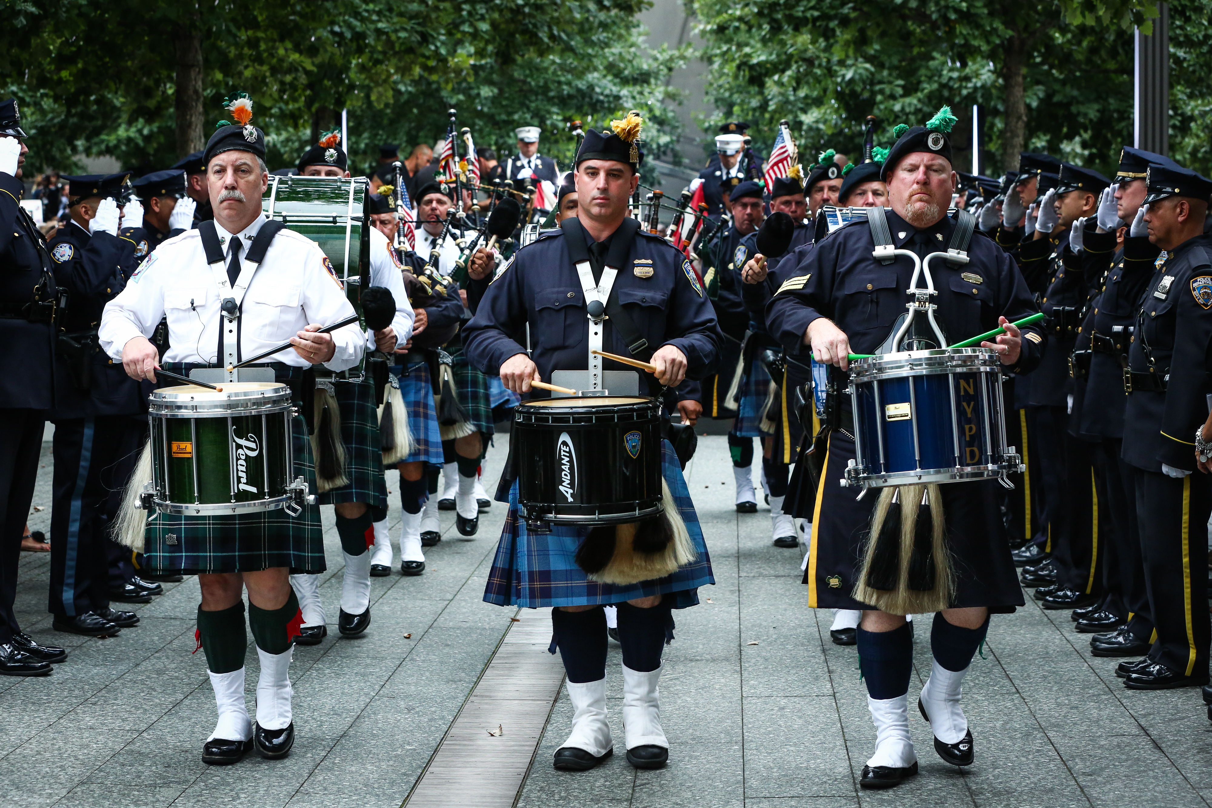 Members of the FDNY, NYPD, and PAPD pipes and drums bands play on Memorial plaza. Three band members in the foreground are wearing kilts and playing snare drums. The band members are lined by uniformed officers who are saluting as the band marches by.