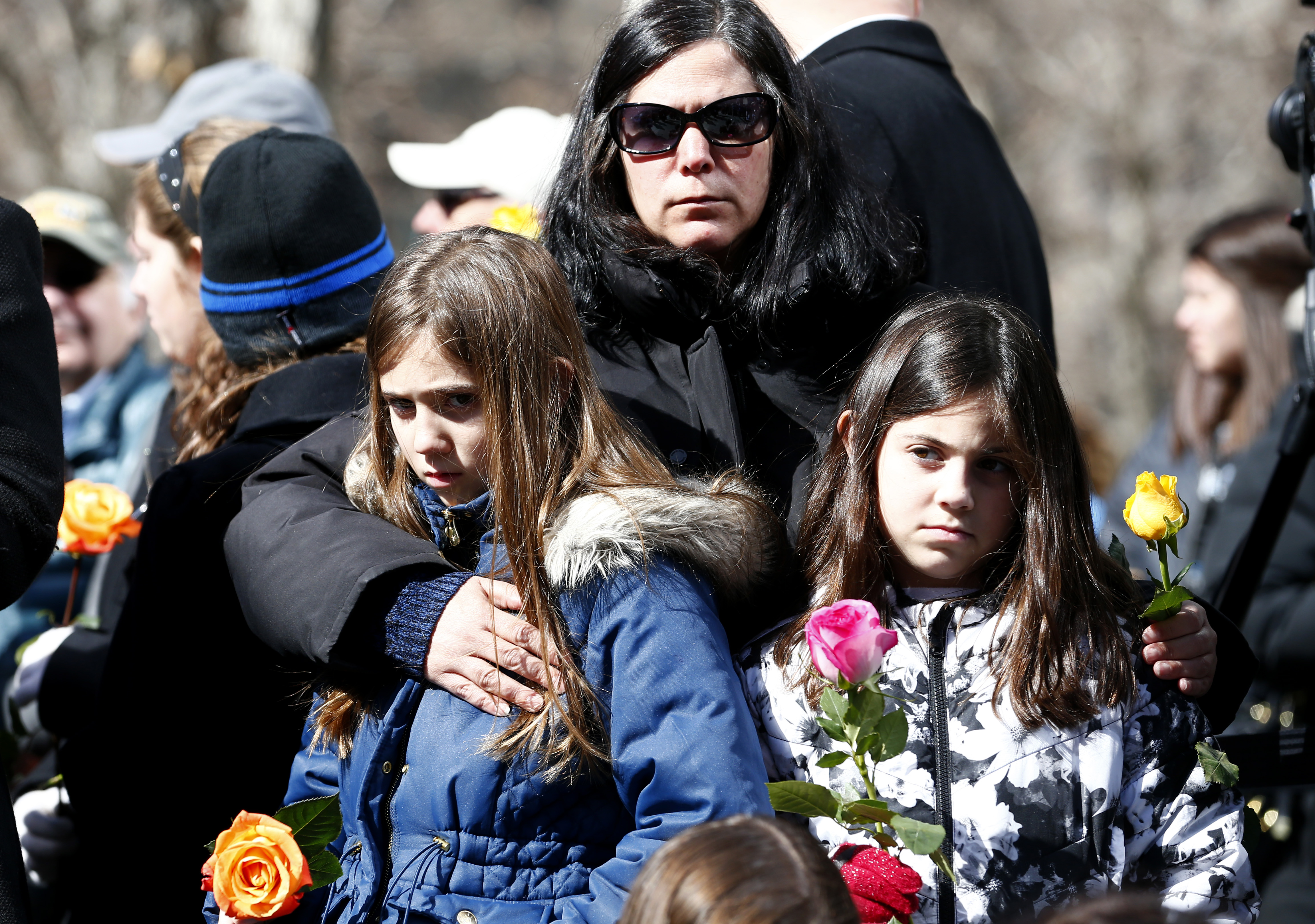 A woman embraces two girls as they stand during a moment of silence on Memorial plaza. The three of them are holding multicolored roses.