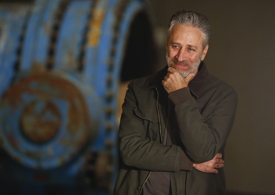 Jon Stewart smiles and rests his head on his hand as he stands at the Museum.