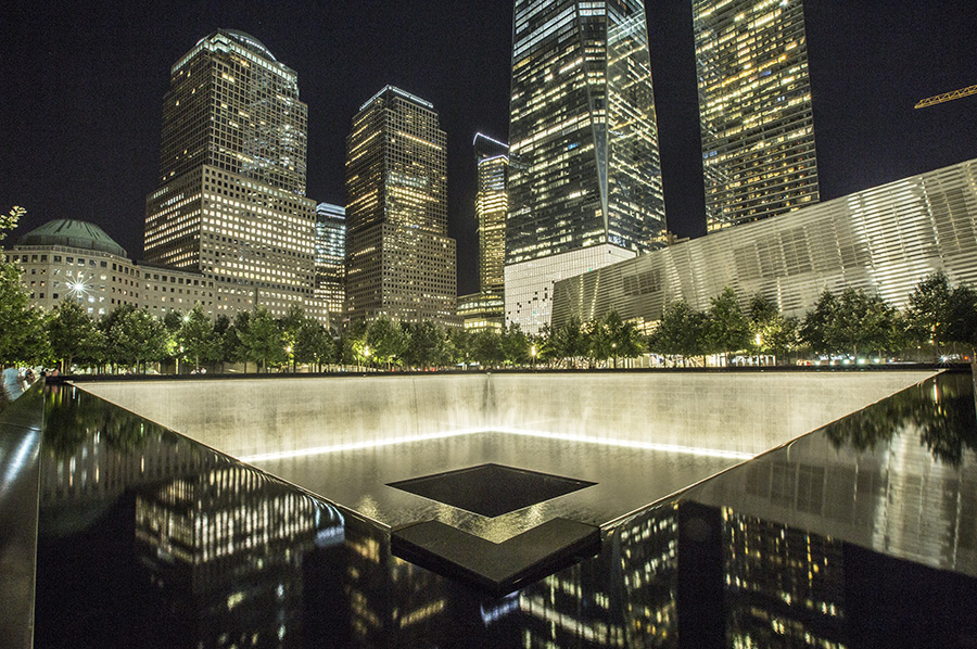 The south pool of the Memorial is lit up at night. The lights of One World Trade Center and nearby buildings reflect in the pool.
