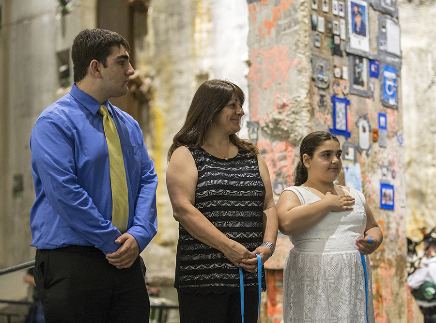 Three members of the Cohen family—a young man, a middle-aged woman, and a girl stand side by side in Foundation Hall. They are holding blue ribbons and standing beside the Last Column to mark the 14th anniversary of the formal end of recovery operations at the World Trade Center.