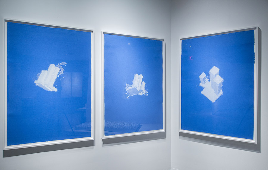 Three works from artist and sculptor Christopher Saucedo’s “World Trade Center as a Cloud” series are framed and displayed on a wall. The works are blue and white and were created by pressing wet layers of white linen with isometric drawings of the World Trade Center onto handmade blue-pigmented cotton pulp.