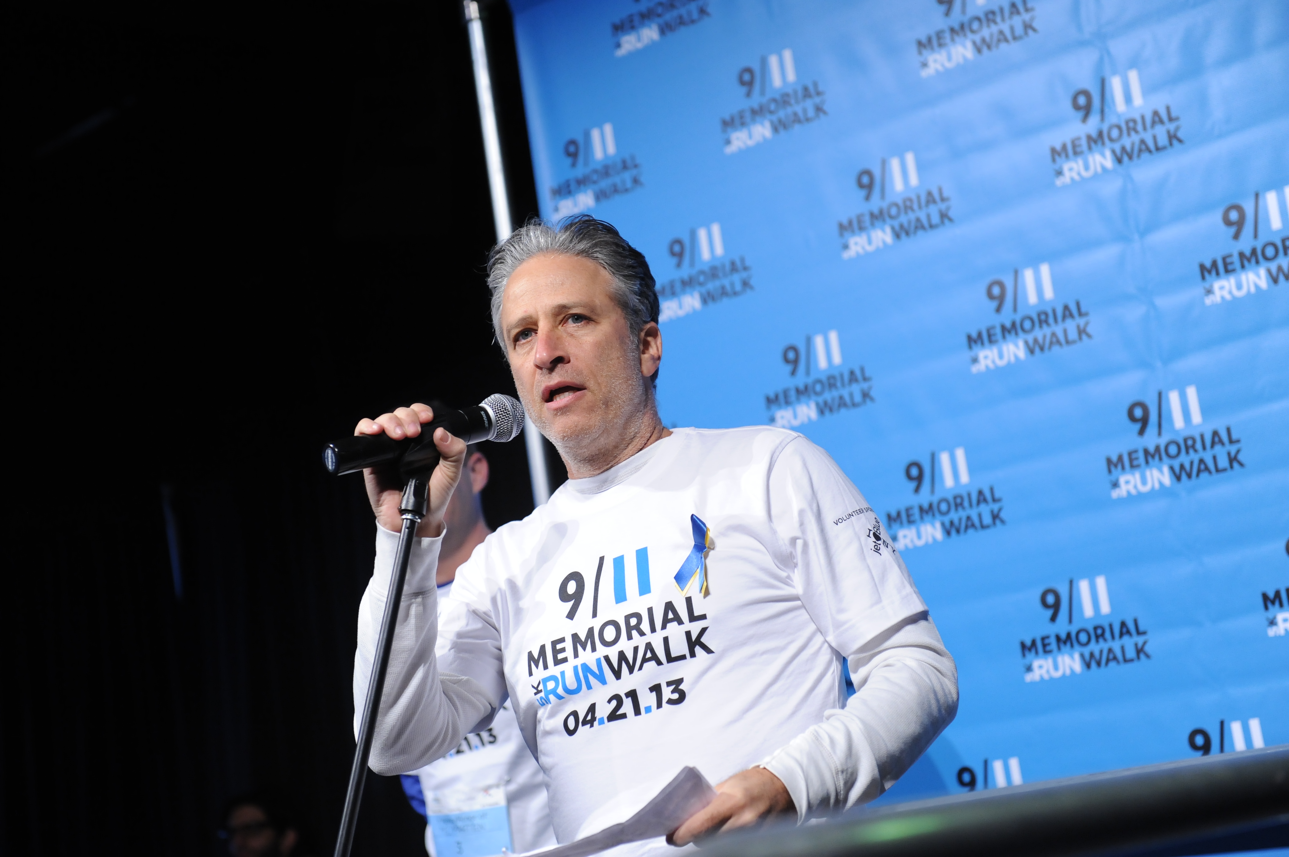 Jon Stewart speaks at a microphone while wearing a 9/11 Memorial 5K Run and Walk shirt.