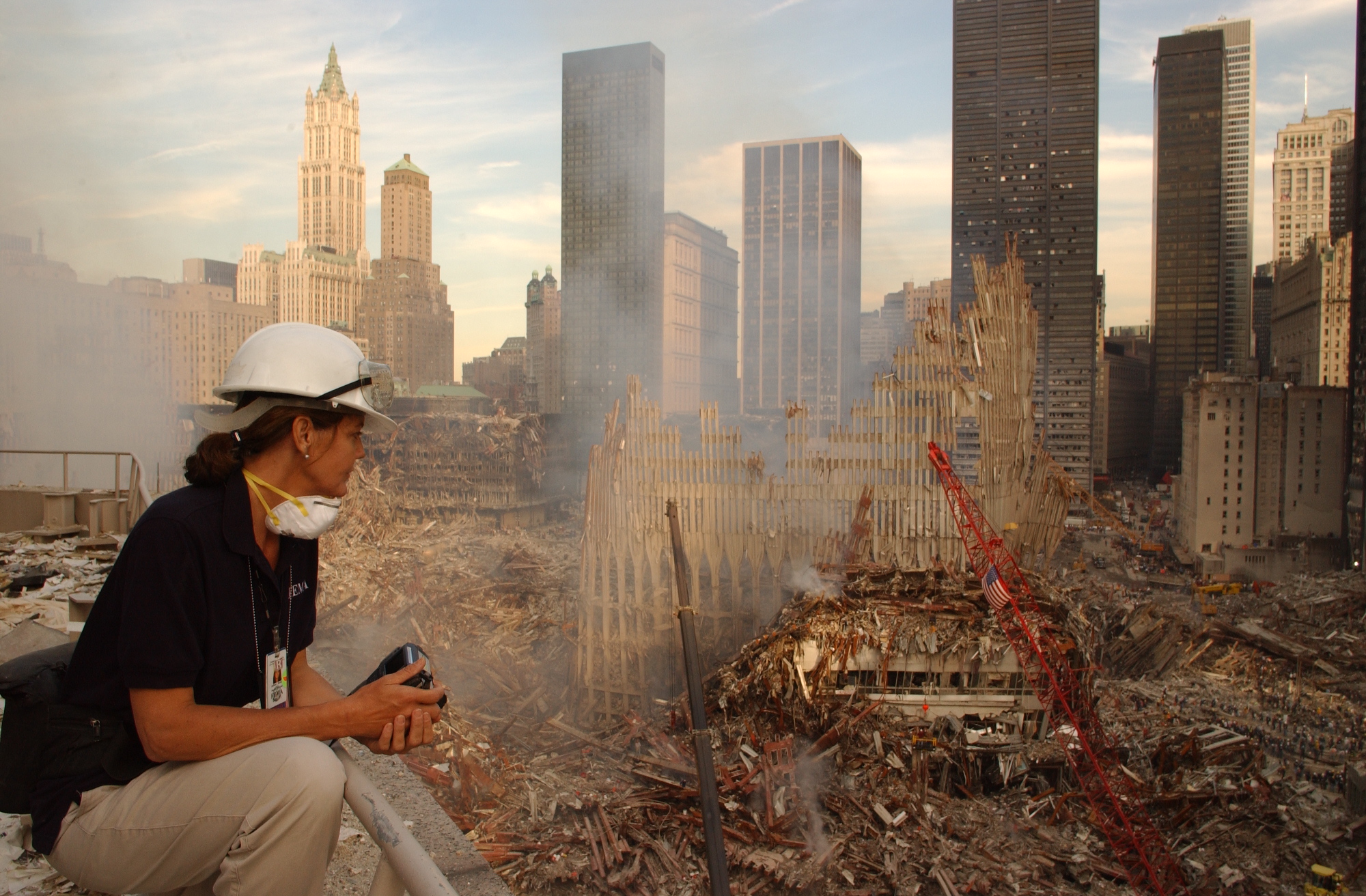 FEMA photographer Andrea Booher looks down at smoking piles of debris at Ground Zero on an evening shortly after the 9/11 attacks. She is wearing a hardhat and a mask as she holds a camera in her hands. The skyscrapers of lower Manhattan are in the distance. Two cranes are in the foreground.