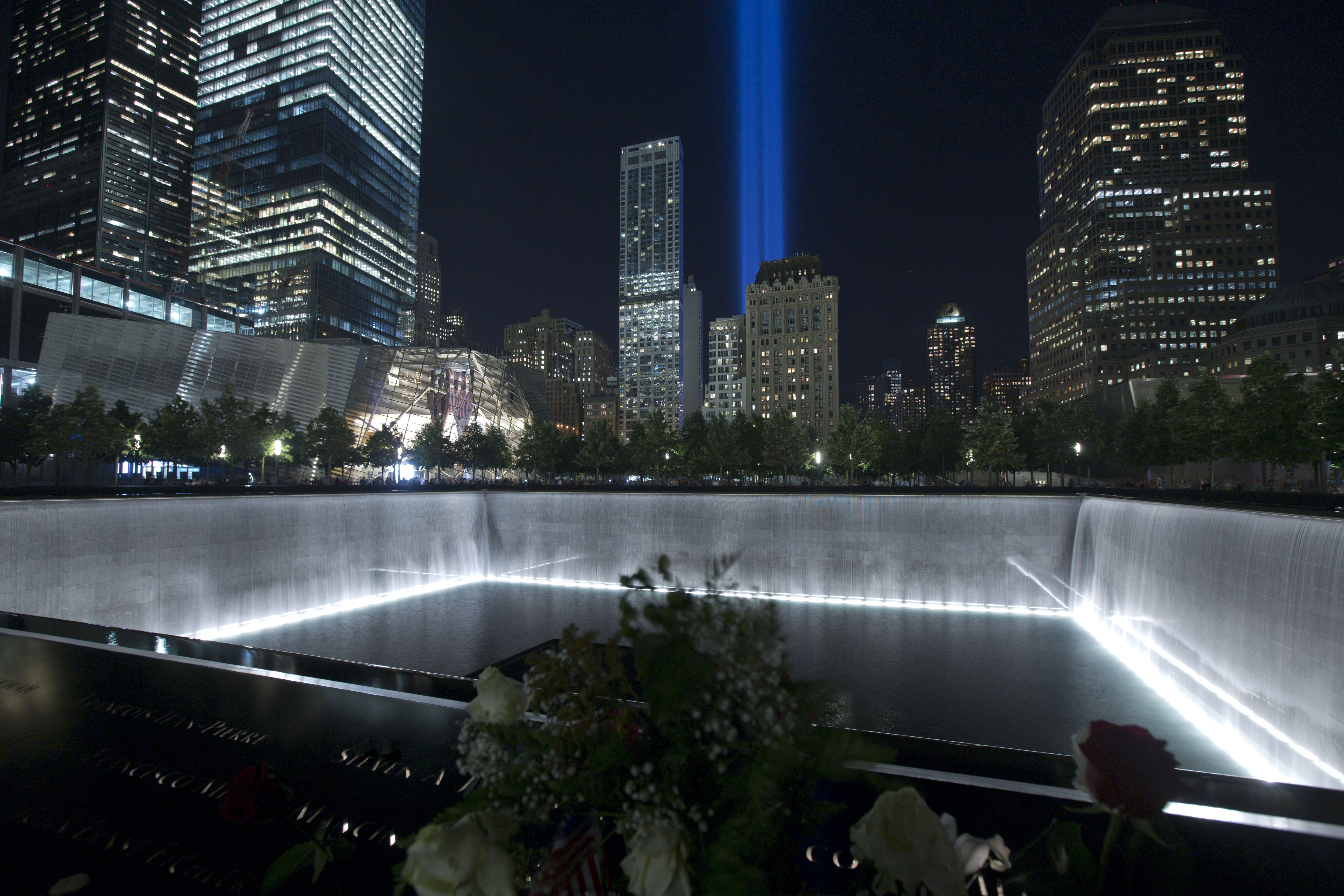 The Tribute in Light shines over the 9/11 Memorial plaza as water cascades down the north reflecting pool. Flowers placed at names on the Memorial’s bronze parapets are in the foreground.