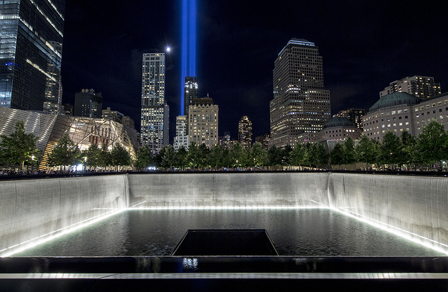 The Tribute in Light is seen shining above Manhattan in this night view of the 9/11 Memorial. The moon glows beside the two blue-white beams symbolizing the Twin Towers. A sprawling reflecting pool is lit up white in the foreground.