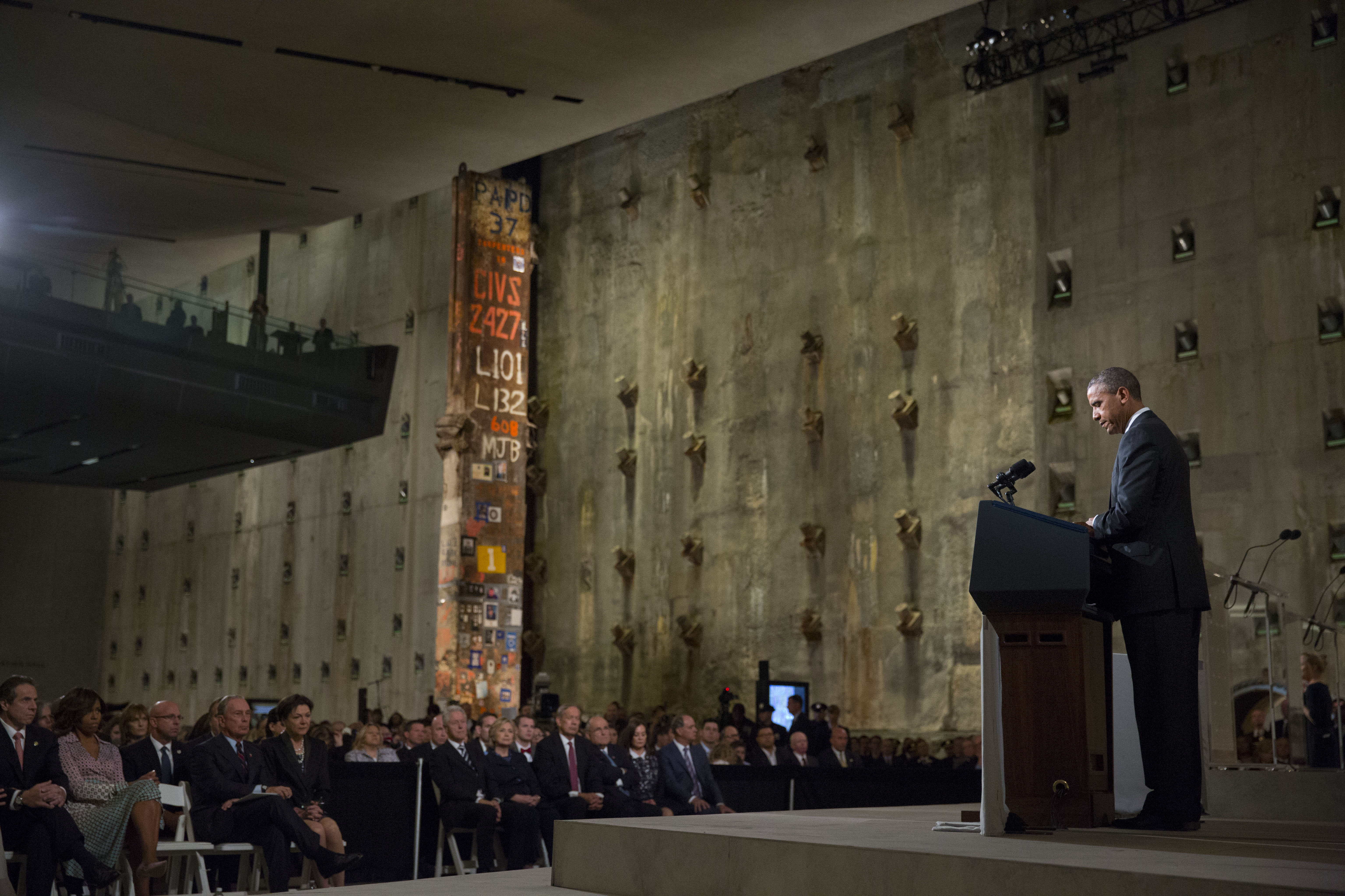 President Barack Obama speaks at a podium during the 9/11 Memorial Museum dedication ceremony. Audience members watch him beside the slurry wall and Last Column.