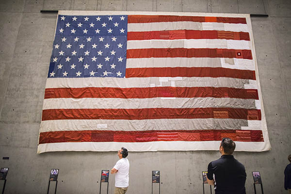 The large national 9/11 flag is displayed on a concrete wall in the south corridor of the 9/11 Memorial Museum. Two visitors are looking up at the flag.