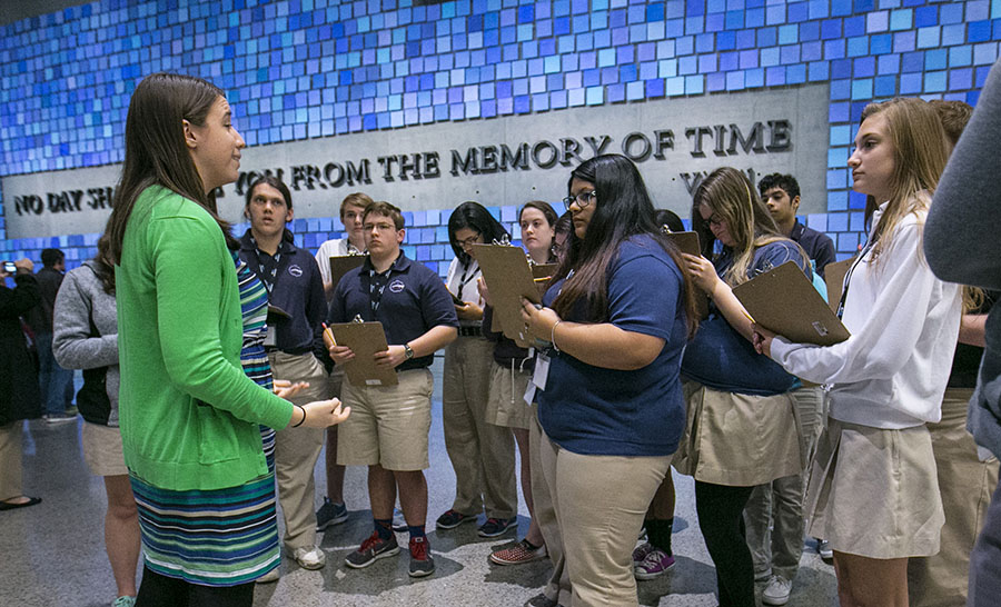 Shannon Elliott, a 9/11 Memorial education specialist, gives a Museum tour to students in Memorial Hall. The students are holding clipboards as they listen to her.