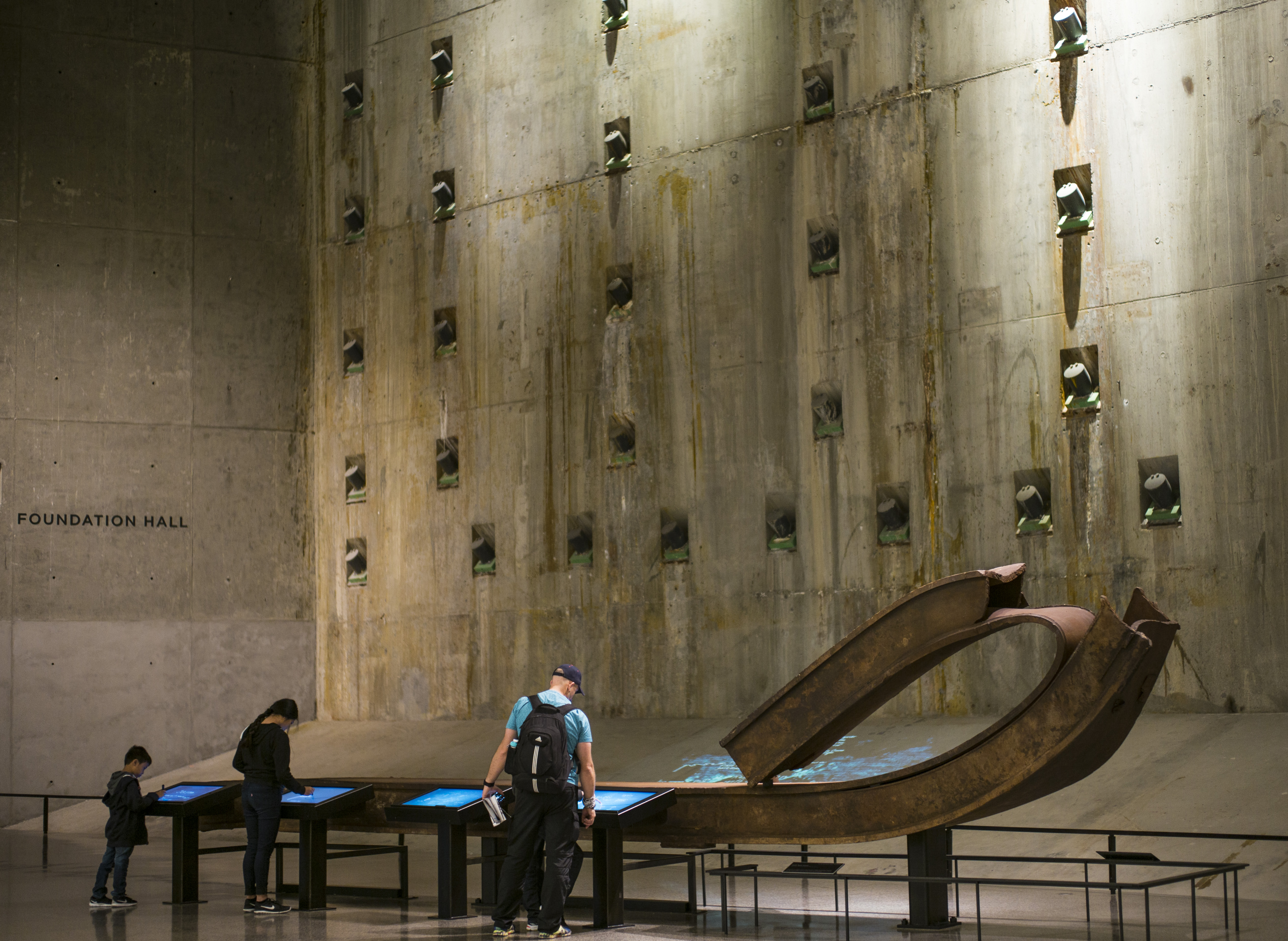 A man, woman boy stand at interactive screens beside a steel beam in Foundation Hall. The slurry wall towers above them.