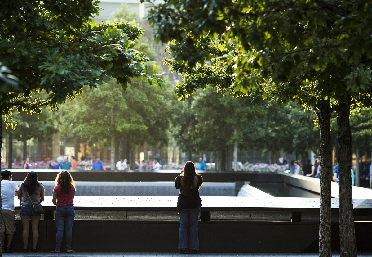 A woman stands alone as she looks out over the North Pool at the 9/11 Memorial on a summer day. Several other visitors are standing off to her left looking out at the pool. And more visitors can be seen across the pool in the distance.