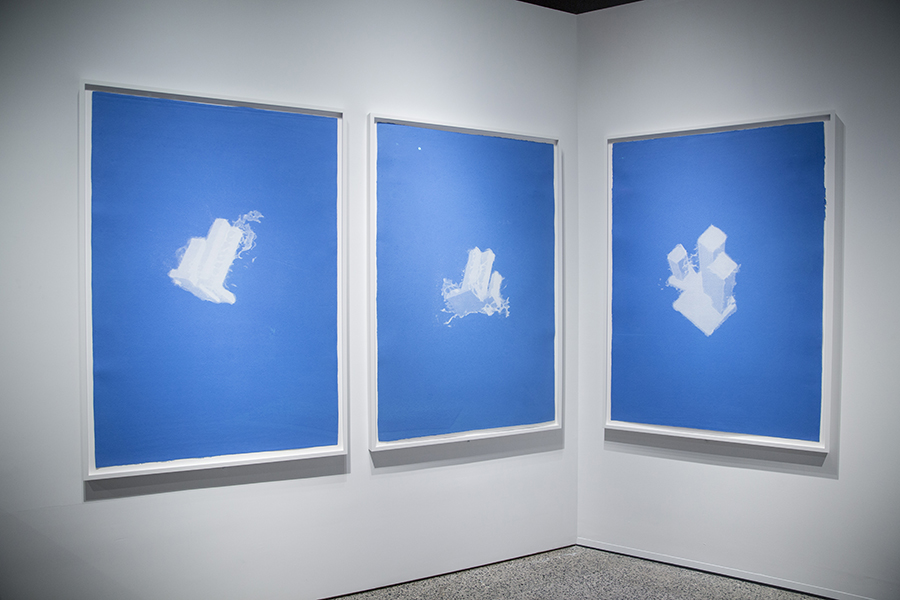 A series of paintings titled “World Trade Center as a Cloud” by artist Christopher Saucedo hang on a white wall at the 9/11 Memorial Museum. The three paintings cloud-like images of World Trade Center buildings floating in a blue sky.