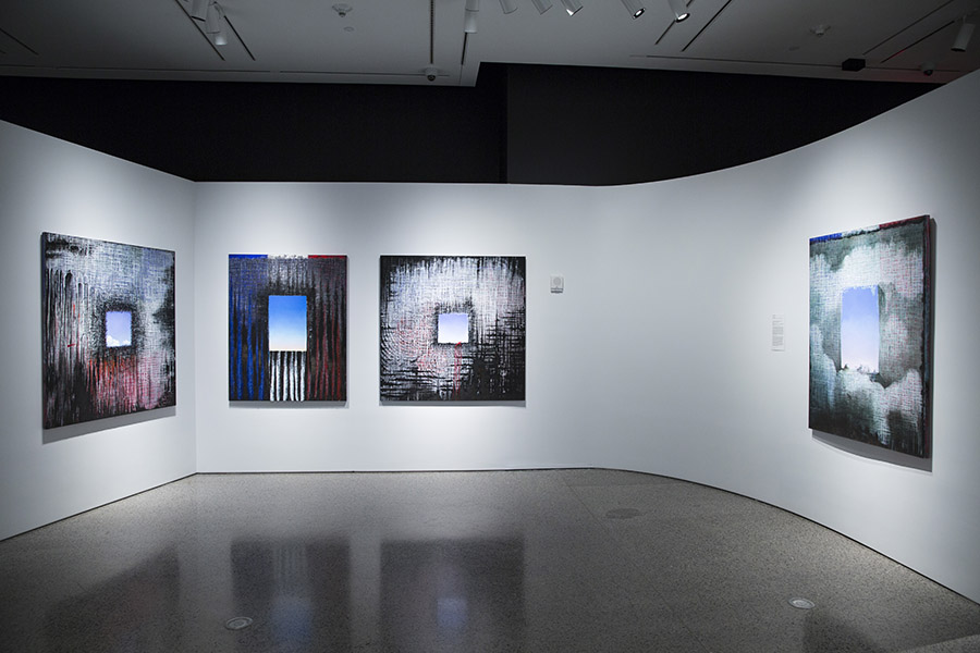 Four of Ejay Weiss’ paintings are seen on display at the 9/11 Memorial Museim. They are a part of his 9/11 Elegies series. The paintings are on square-shaped canvases. Square-shaped holes painted the color of  the sky are at the center. These squares are surrounded by various abstract designs in black, white, red, and blue.