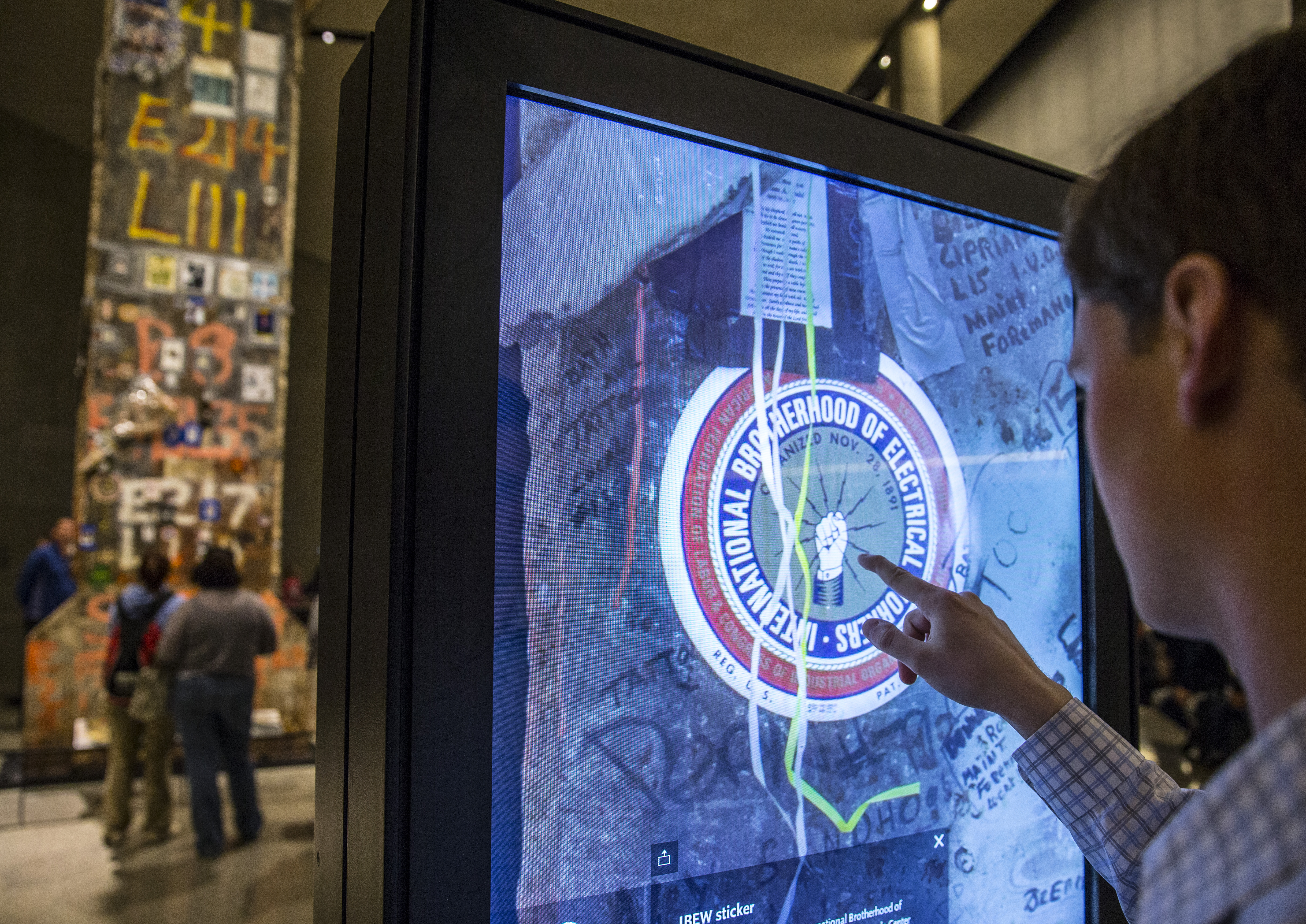 A man in Foundation Hall uses an interactive touchscreen that is displaying a photo of an International Brotherhood of Electrical Workers sticker that is on the Last Column.