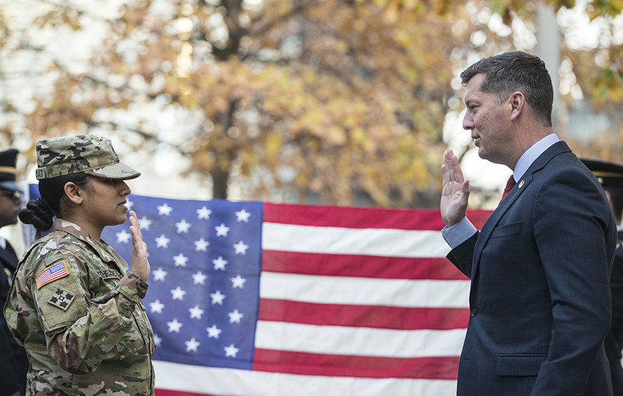 Under Secretary of the Army Patrick Murphy officiates a promotion ceremony on the 9/11 Memorial. He is holding up his right hand as he stands across from a woman in a U.S. military outfit who is also holding up her right hand. An American flag is hanging in the background.